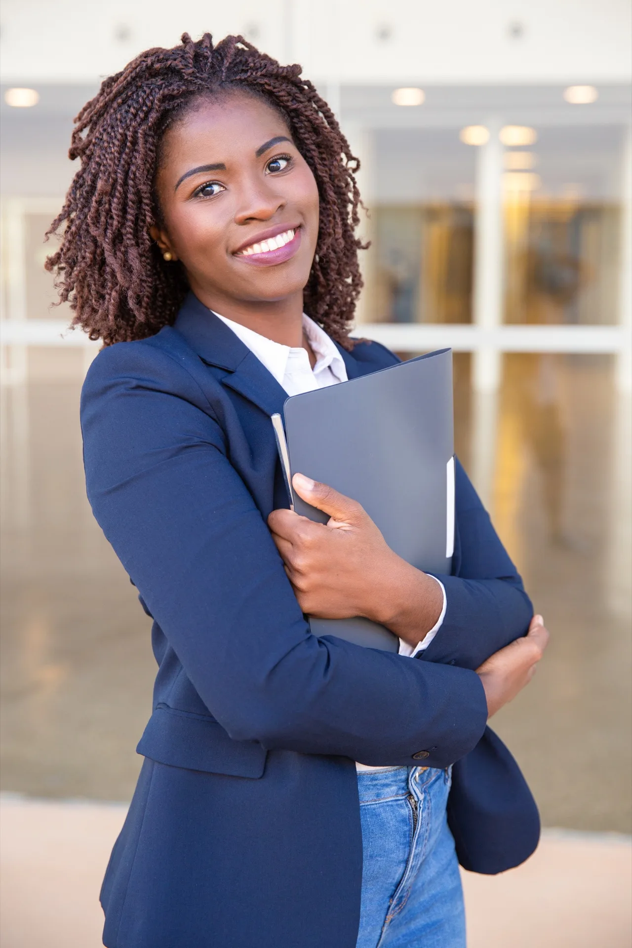 Jeune diplômée africaine souriante tenant un porte-documents devant l'entrée d'un bâtiment universitaire moderne.